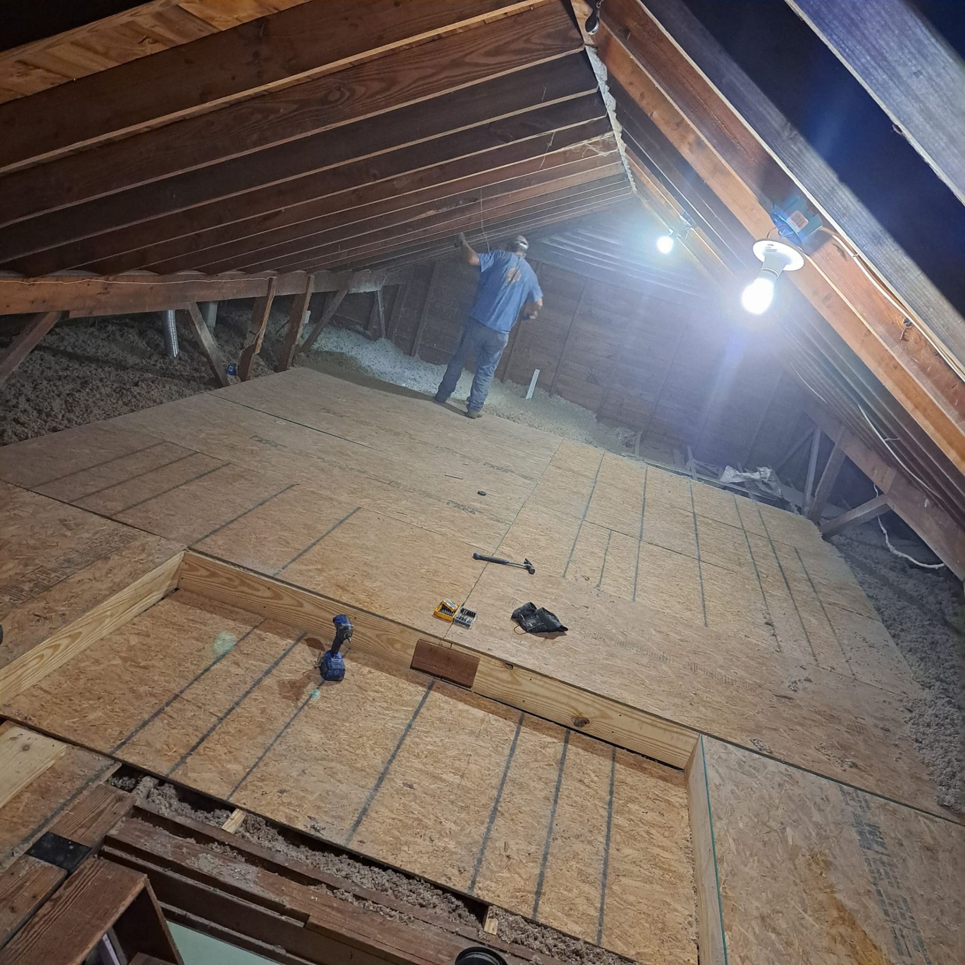 Worker standing on plywood floor in a dim attic under exposed beams and a bright light bulb