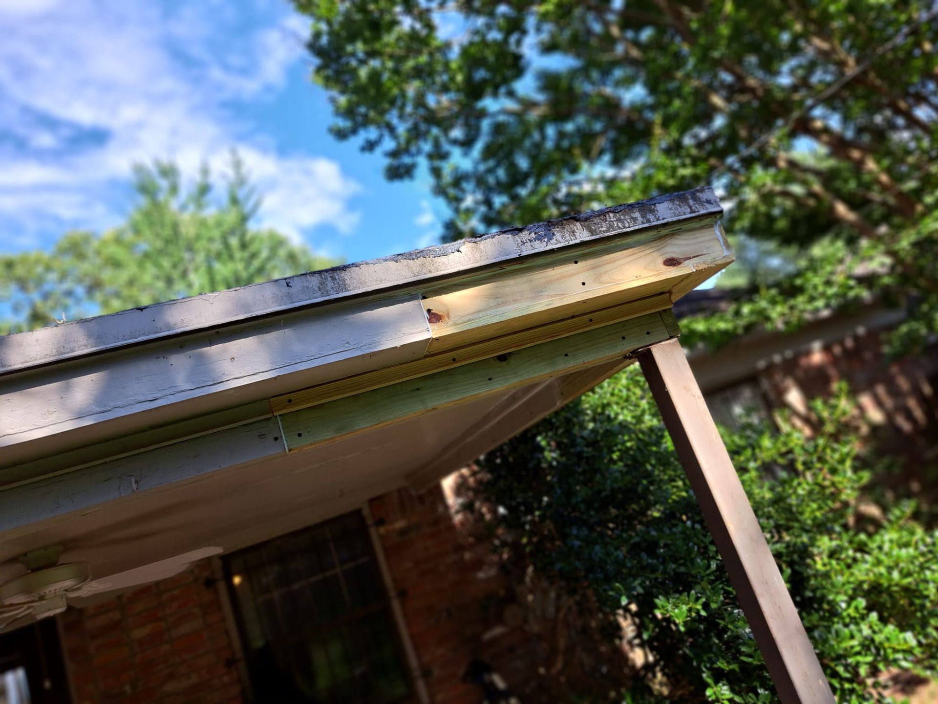 Close-up of a house roof edge and wooden support post against a blue sky and trees