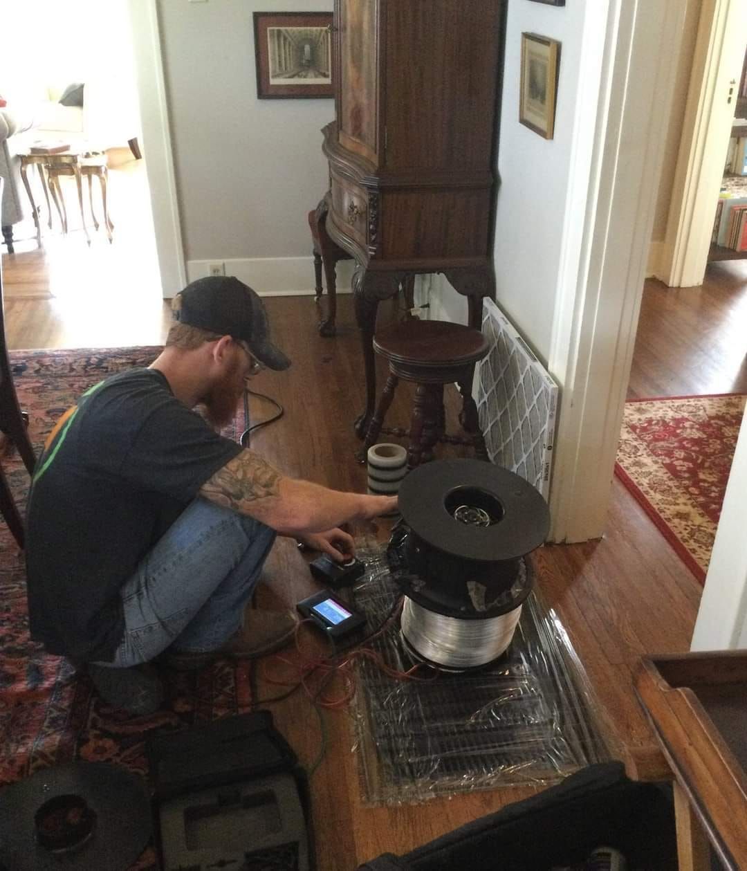 Man in a black cap setting up a small black heater on a hardwood floor in a living room