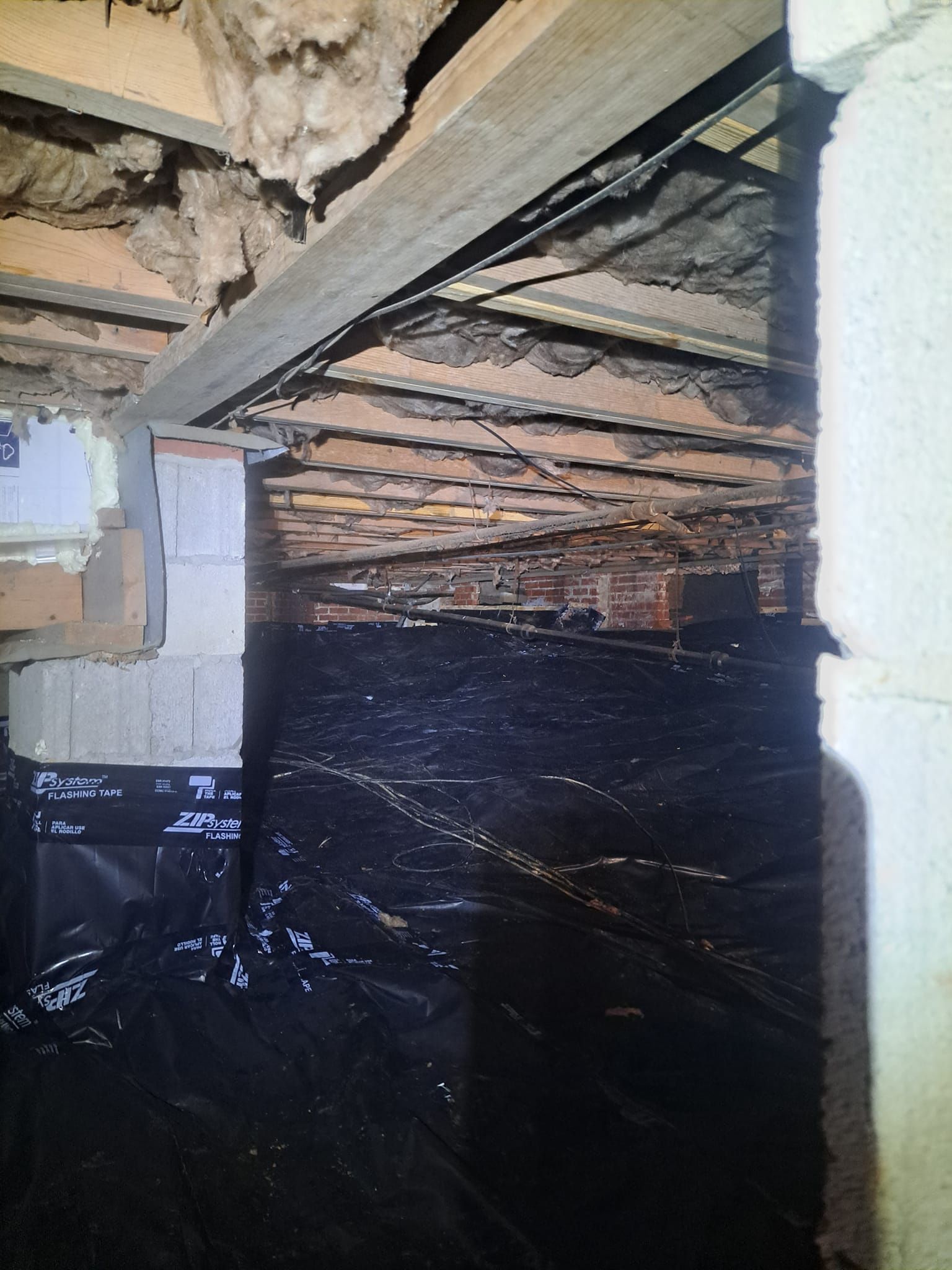 Crawlspace under a house with black plastic vapor barrier and exposed wooden floor joists above