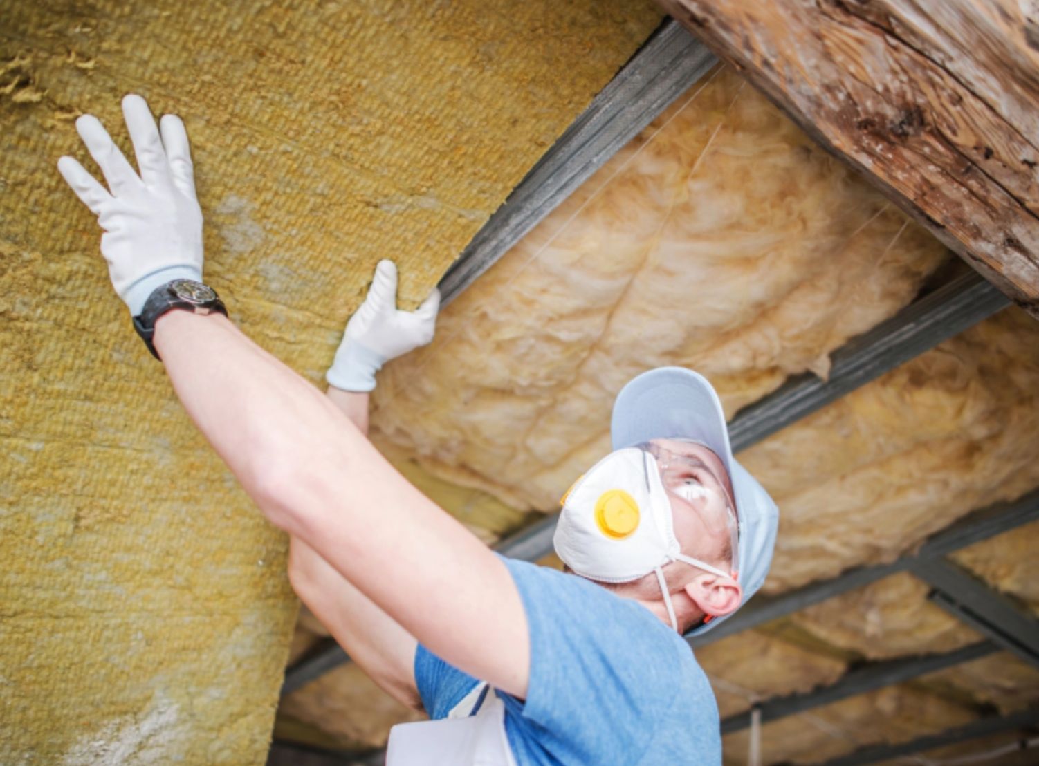 Worker in gloves and mask installing yellow insulation in a ceiling cavity