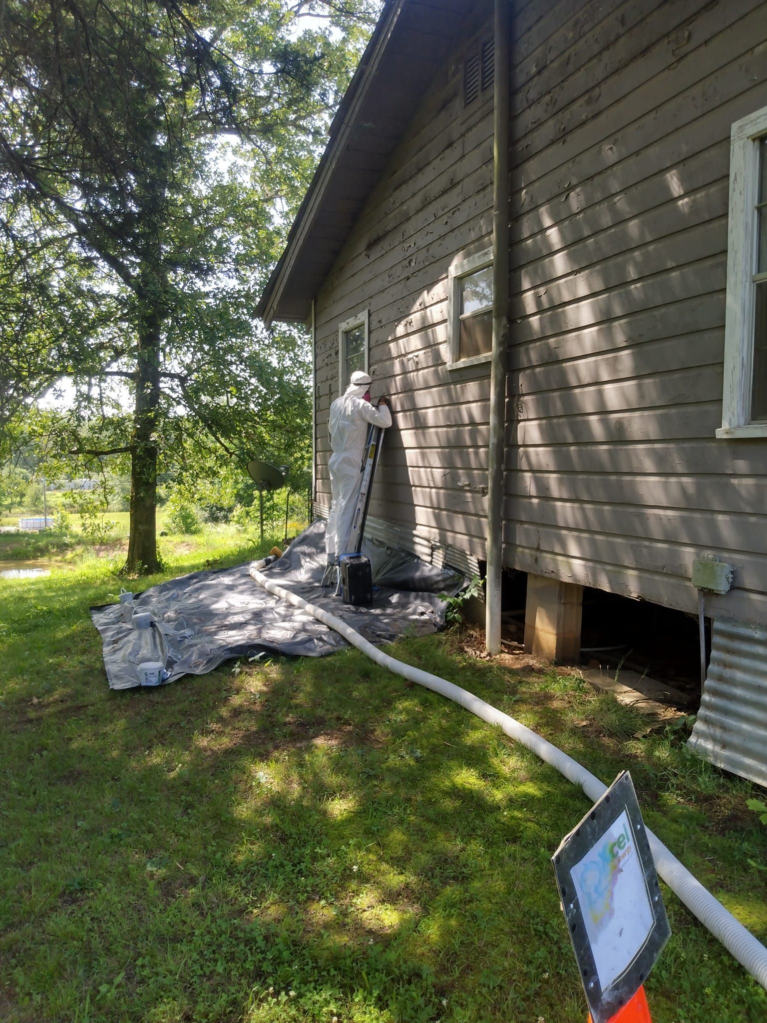 Person spray-painting or coating the side of a house beside a drop cloth and hose in a sunny yard