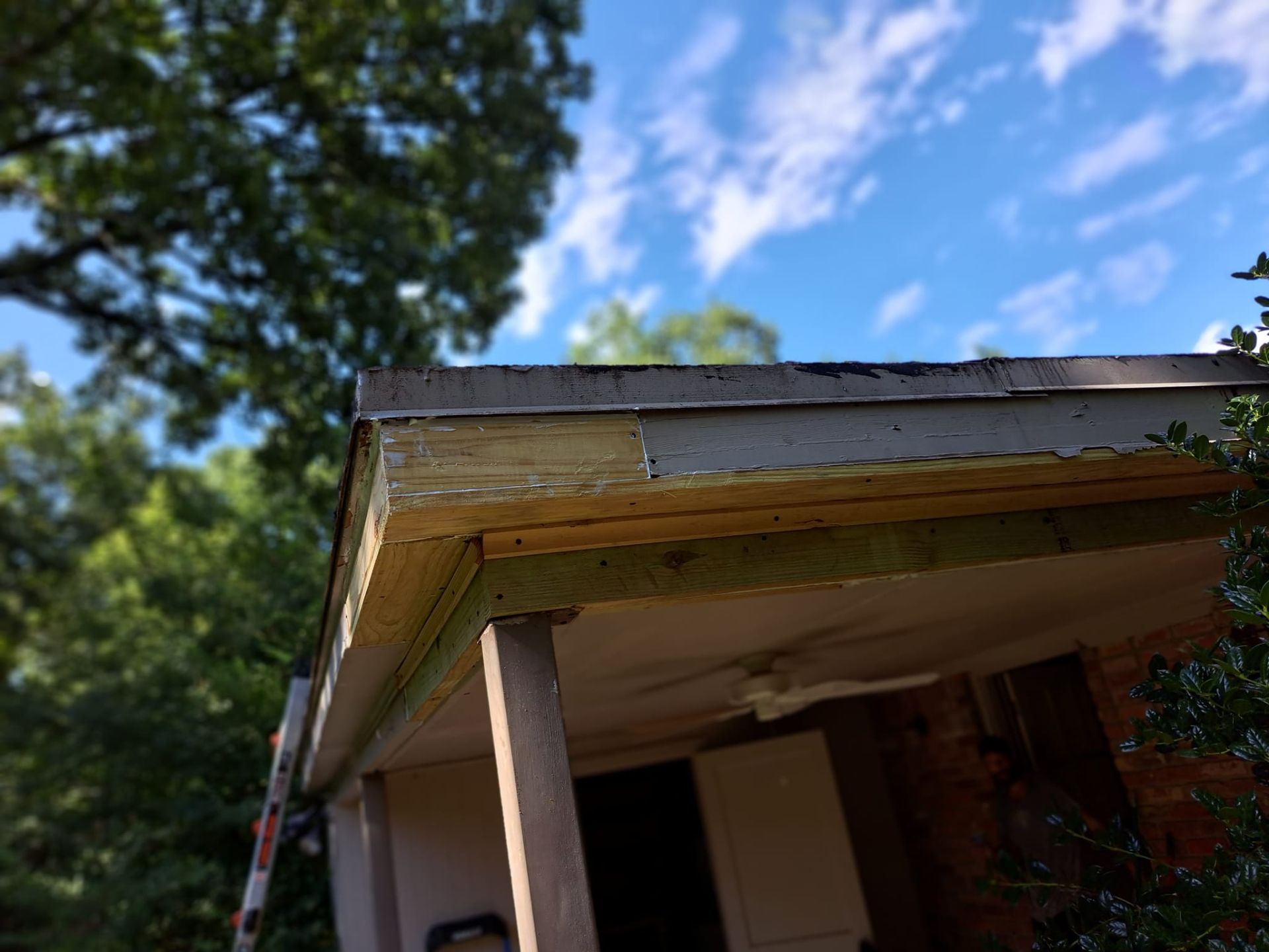 Damaged porch roof with exposed wood and a ceiling fan, viewed from below against a blue sky.