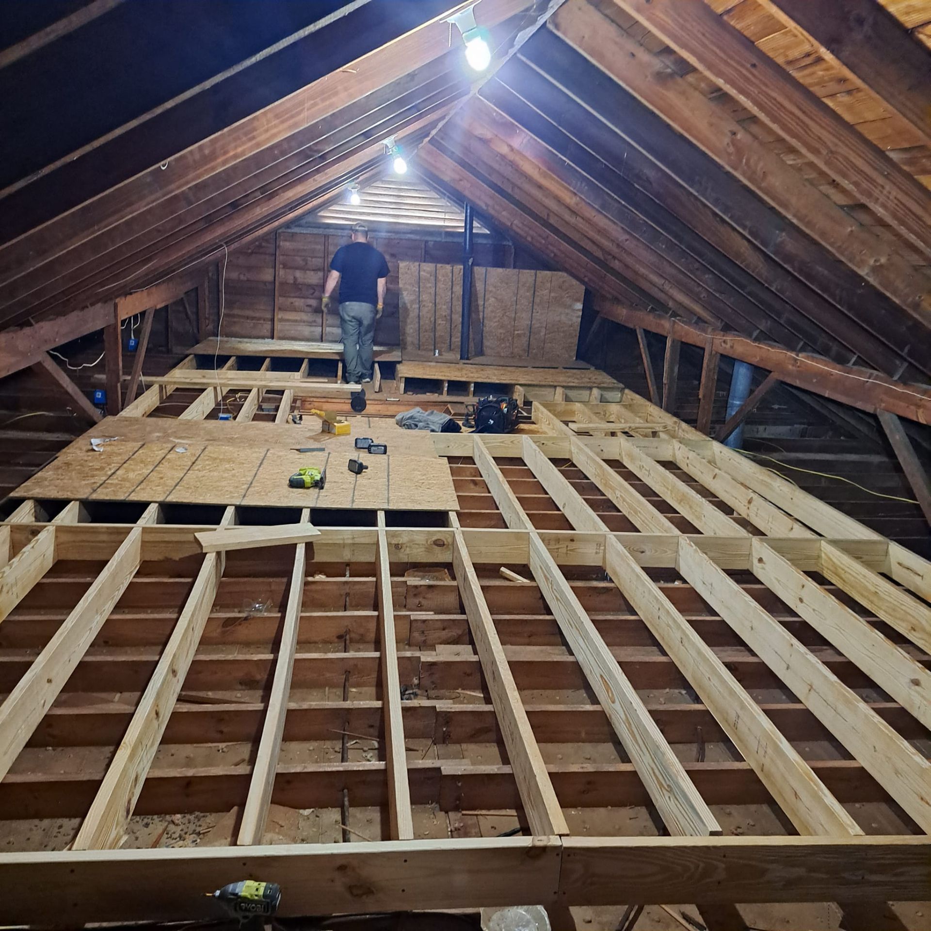 Attic with exposed rafters and new floor joists under construction, with a worker in the distance.