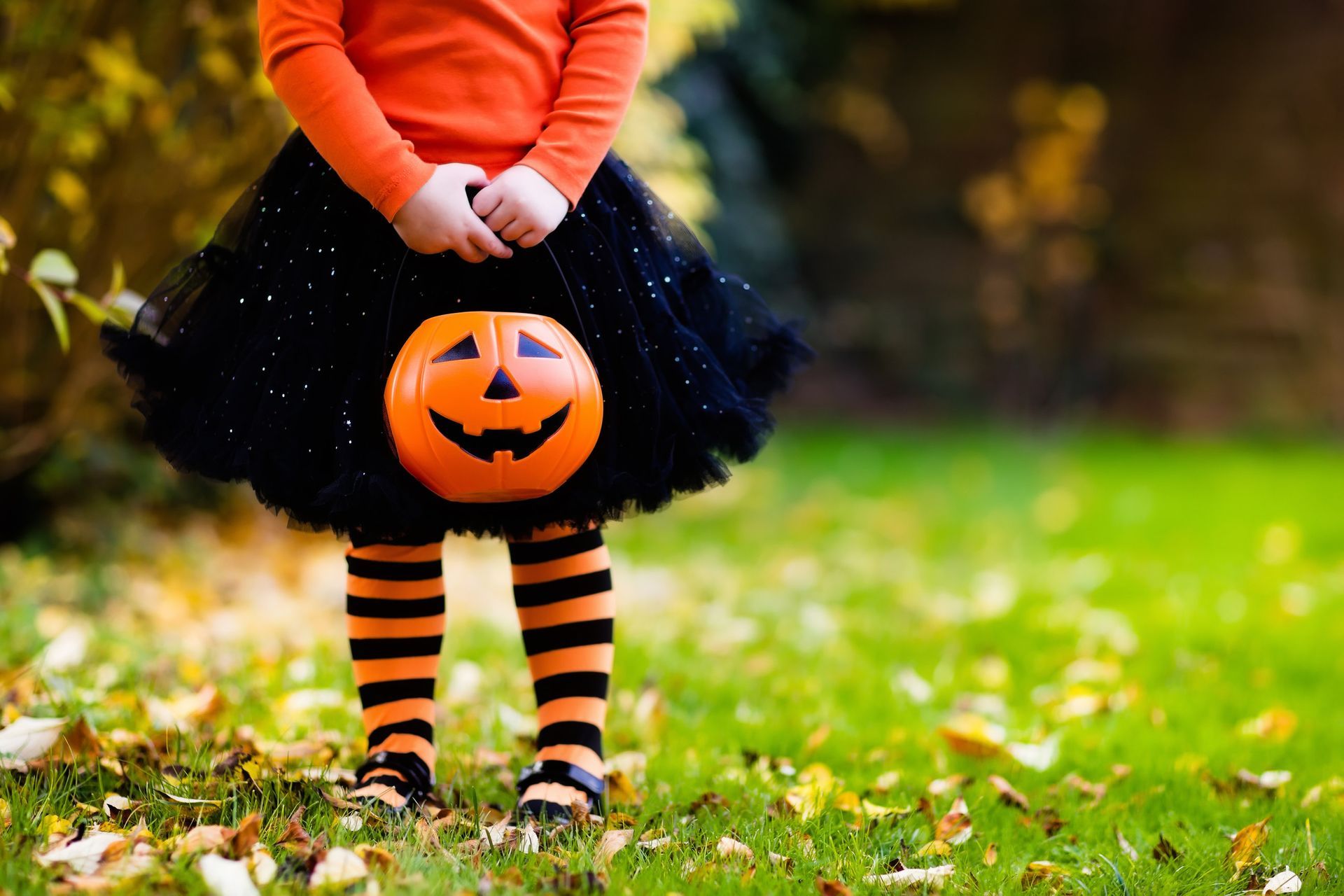 Child in orange sweater and black tutu with jack-o'-lantern face, standing on grass with autumn leaves.