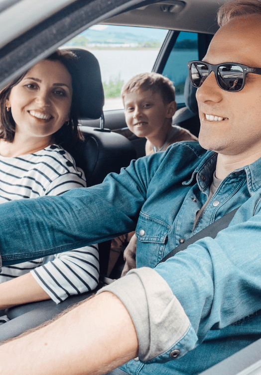Three people smiling in a car, with the driver wearing sunglasses and a denim shirt.