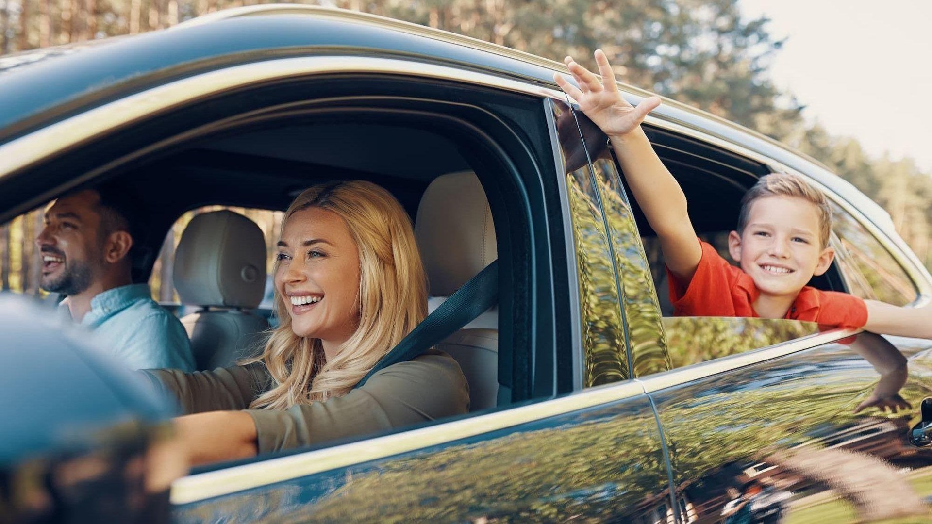 Family smiling in a black car, with a child waving from the back window on a sunny day