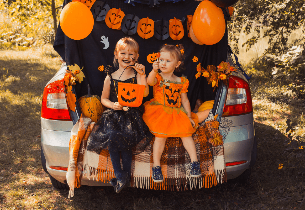 Two children in Halloween costumes sitting in the back of a car decorated with orange and black pumpkins