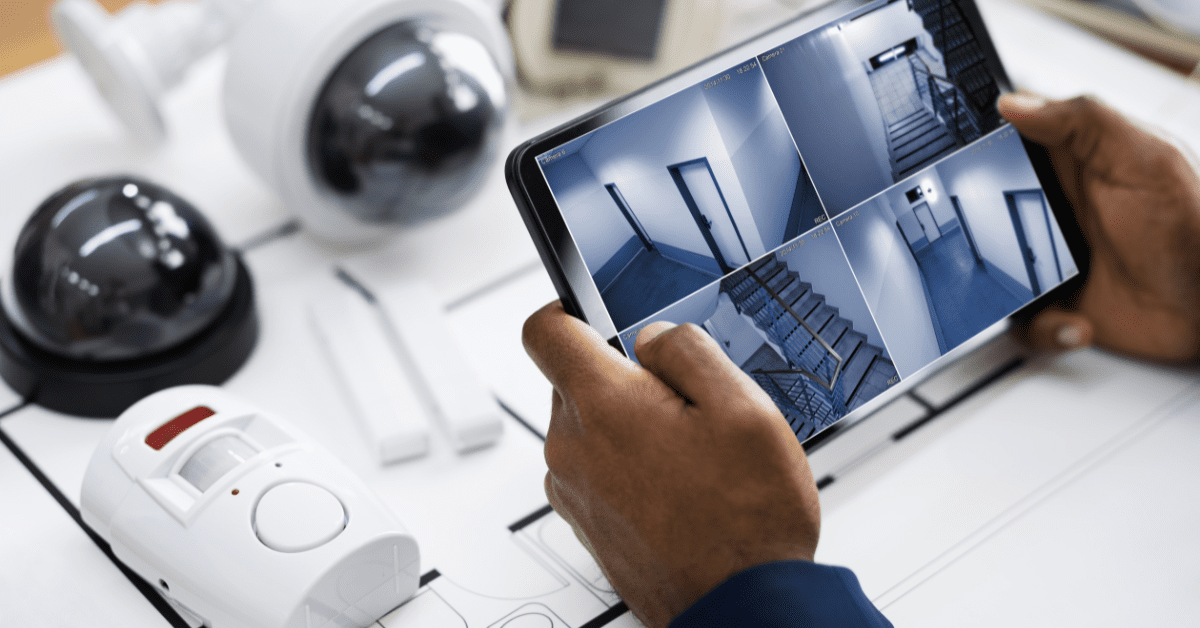 Hands holding a tablet showing a home security camera feed beside several surveillance cameras.