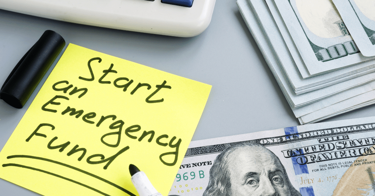 Sticky note saying “Start an Emergency Fund” with cash and a dollar bill on a desk