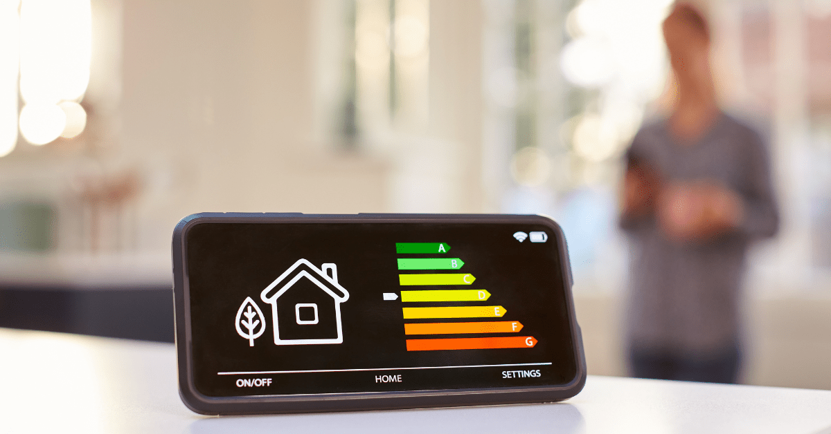 Smartphone showing a home energy-efficiency meter with green-to-red bars on a table, blurred person in background