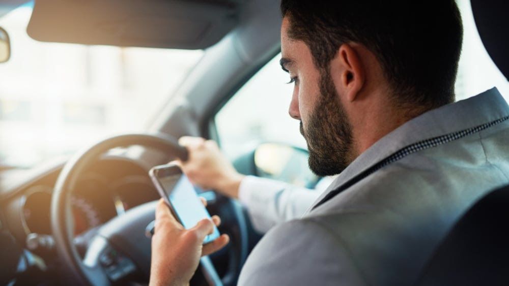 Driver in a car glancing at a smartphone while holding the steering wheel, dashboard visible.