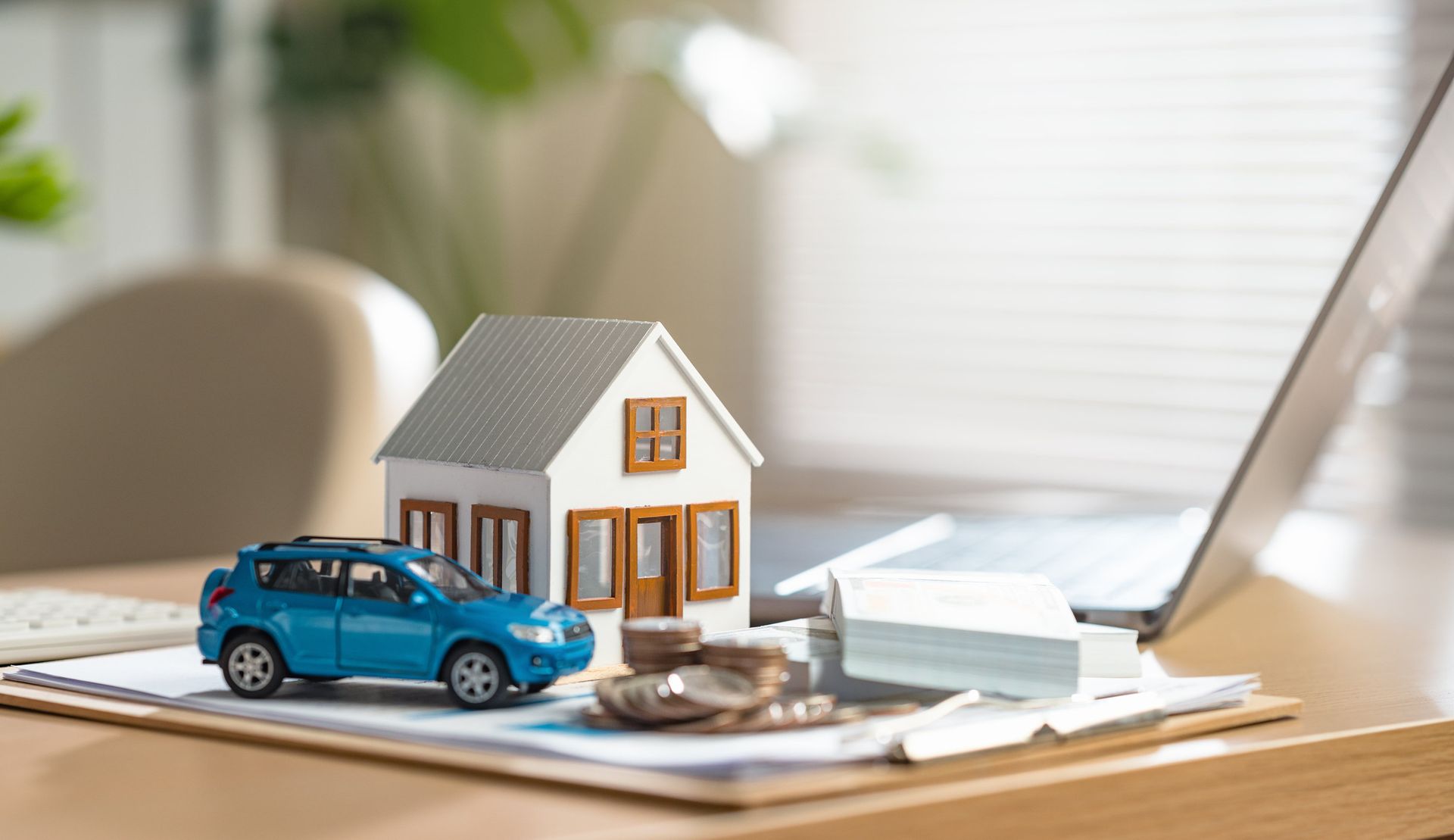 Miniature house model with a blue car and stacks of papers on a desk beside a laptop.