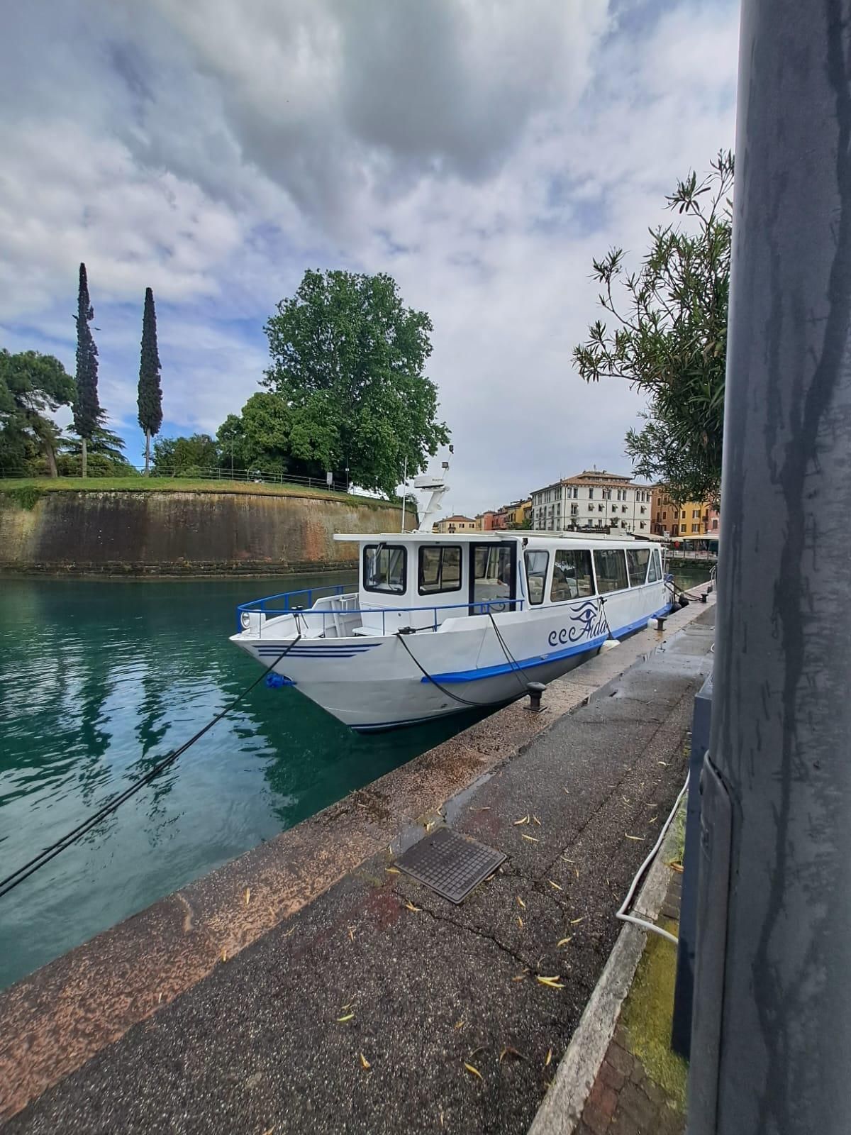 Barca turistica bianca ormeggiata in un canale accanto a un argine di pietra, con alberi ed edifici sotto un cielo nuvoloso.
