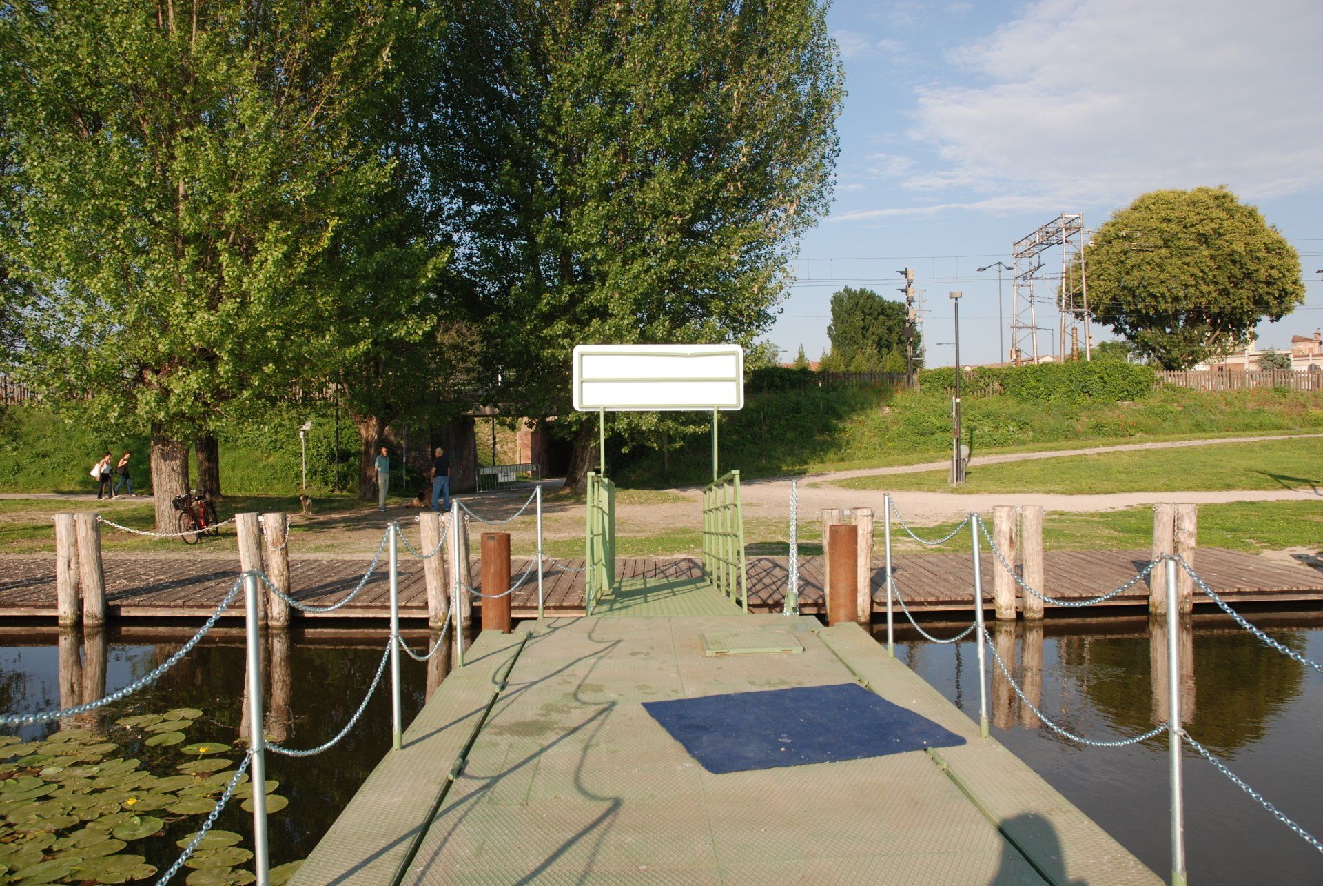 Pontile galleggiante con ringhiere di corda su uno stagno, delimitato da alberi e un cartello vuoto in un parco soleggiato.
