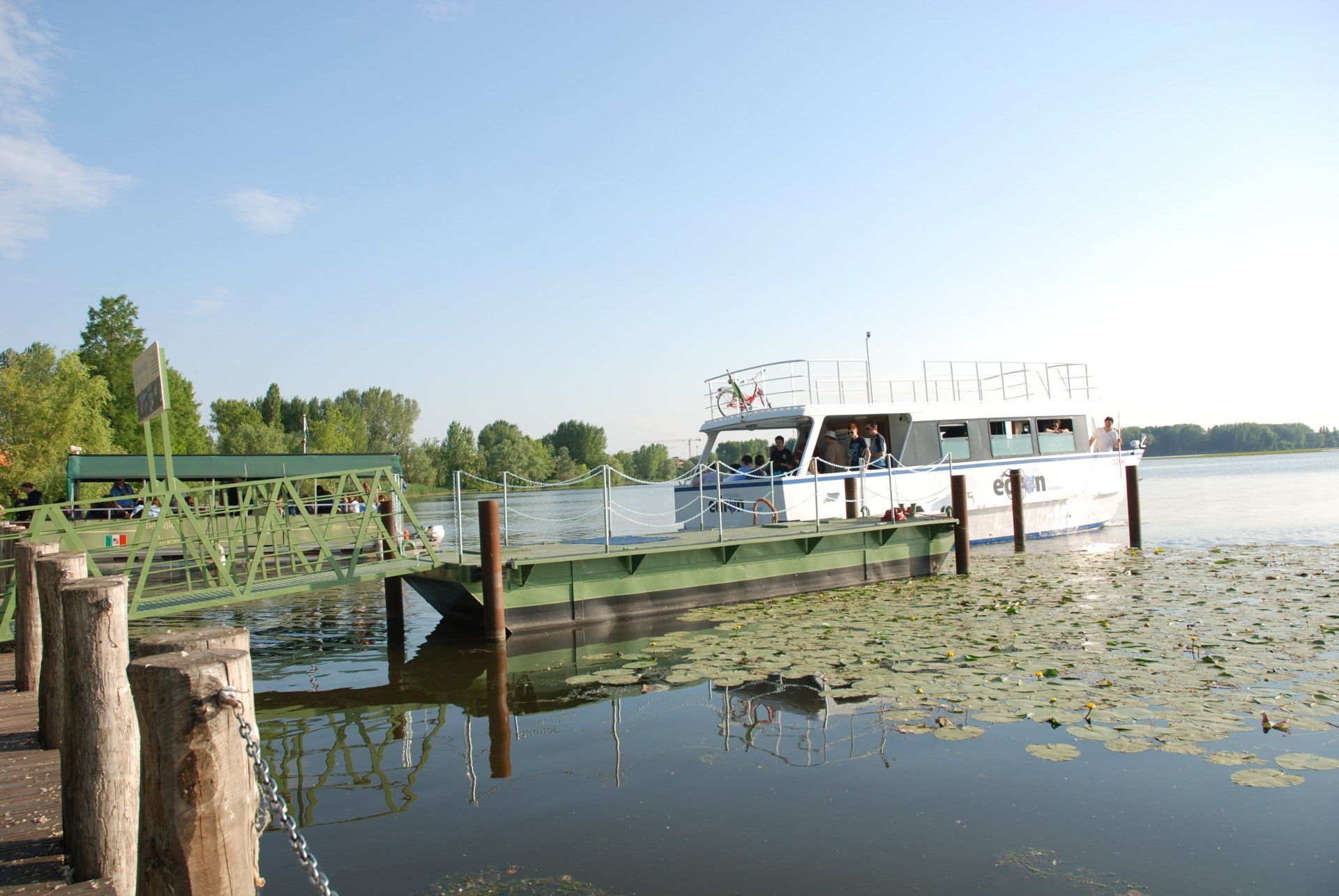 Un traghetto bianco attraccato a un molo su un fiume tranquillo e ricoperto di ninfee, sotto un cielo azzurro e limpido.