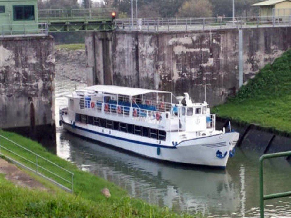 Una barca turistica bianca attraversa una chiusa del canale, incastonata tra muri di cemento e rive verdi.
