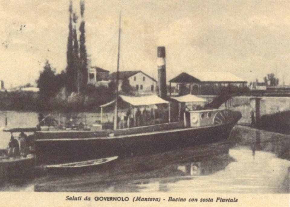 Scena fluviale in tonalità seppia con una lunga imbarcazione ormeggiata accanto a un canale, alberi ed edifici sullo sfondo.