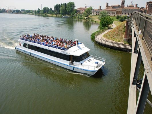 Un battello turistico affollato di passeggeri naviga lungo un fiume costeggiando un ponte e gli edifici che si affacciano sulla riva.