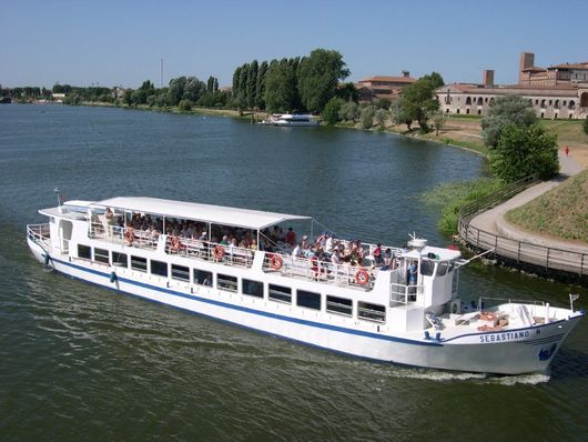 Un battello turistico bianco, carico di passeggeri, naviga su un fiume costeggiato da alberi.