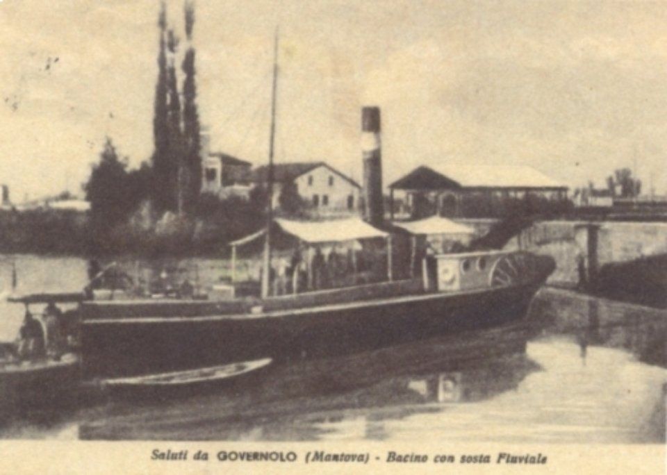 Foto d'epoca in bianco e nero di un battello fluviale ormeggiato vicino a una cittadina sul fiume, con alti alberi ed edifici sullo sfondo.