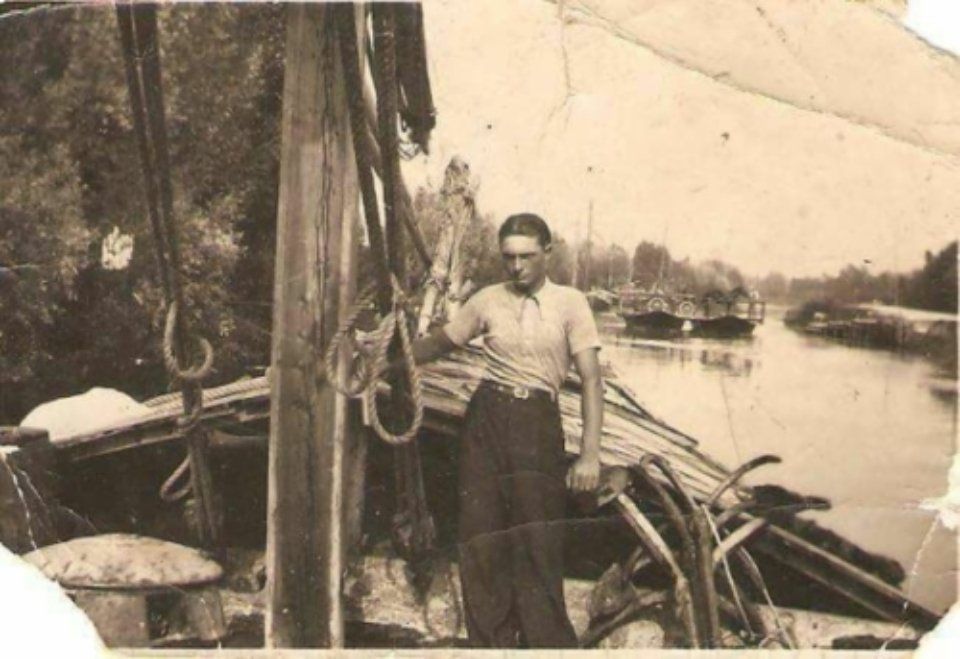 Persona in piedi su una barca di legno in riva a un fiume, con remi e altre imbarcazioni sullo sfondo.