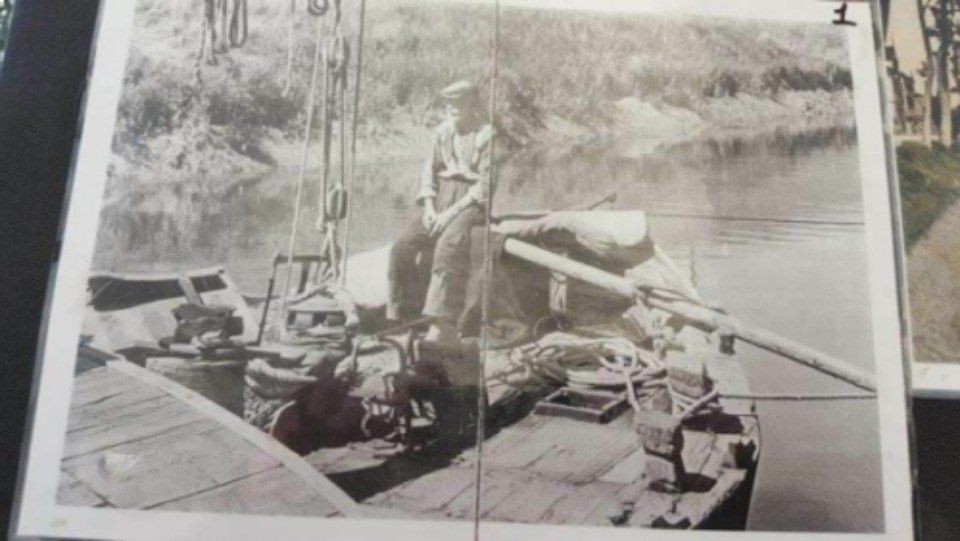 Due persone a bordo di una piccola imbarcazione caricano un tronco su una macchina metallica accanto alla riva di un fiume.