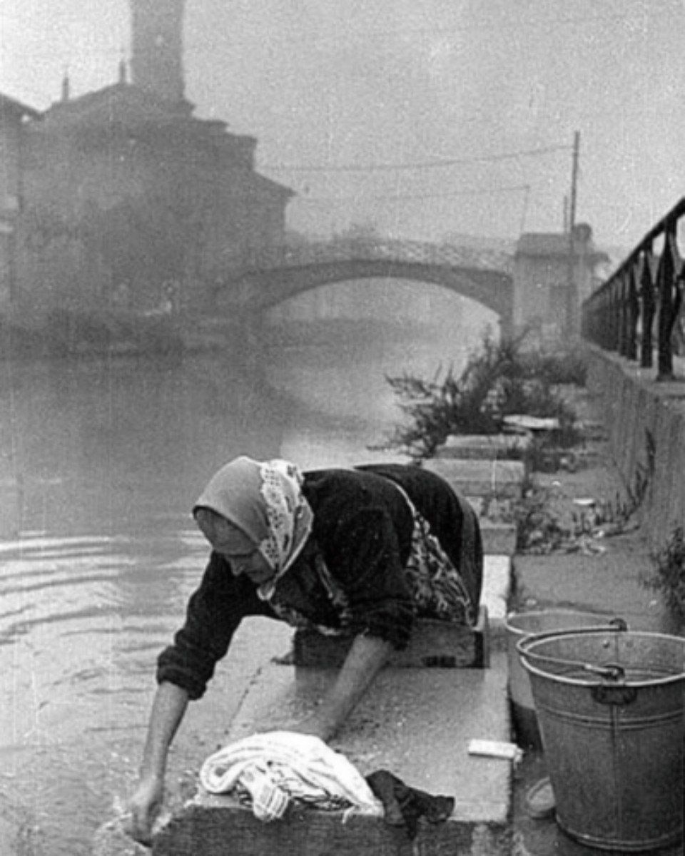 Una donna lava i panni in riva a un fiume, con un ponte e degli edifici sfocati sullo sfondo.