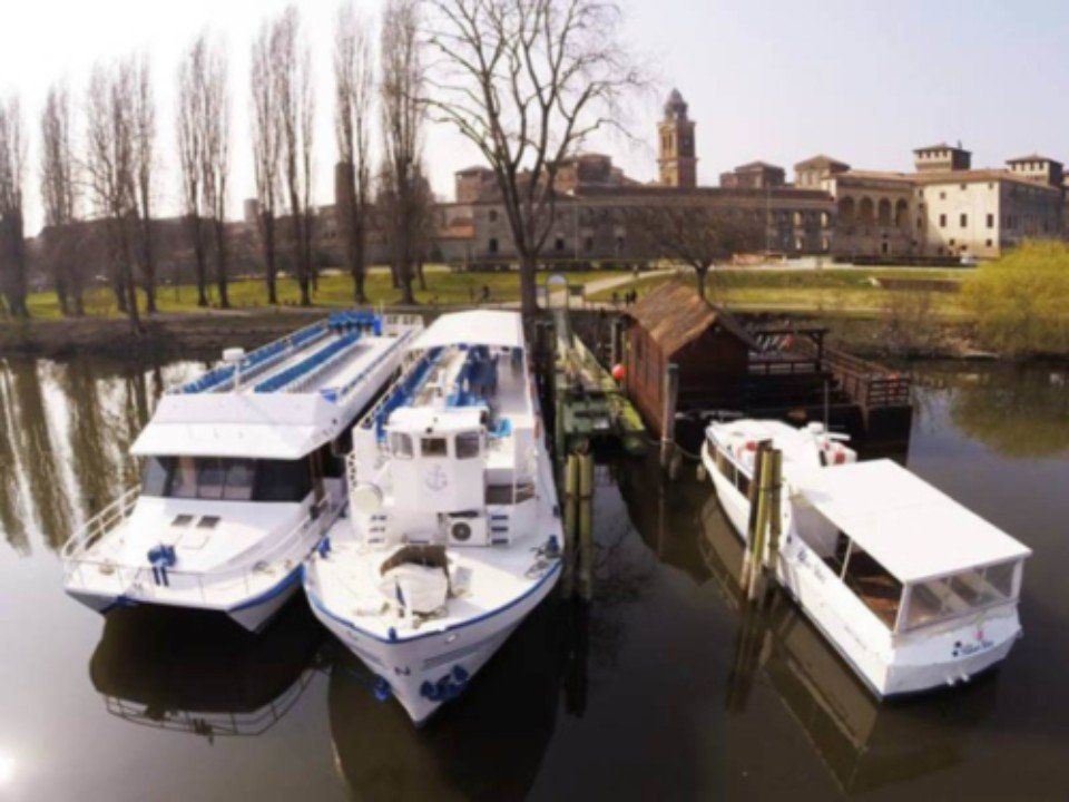 Tre barche ormeggiate su un fiume tranquillo accanto a un piccolo ponte e ad alcuni alberi, con edifici sullo sfondo.