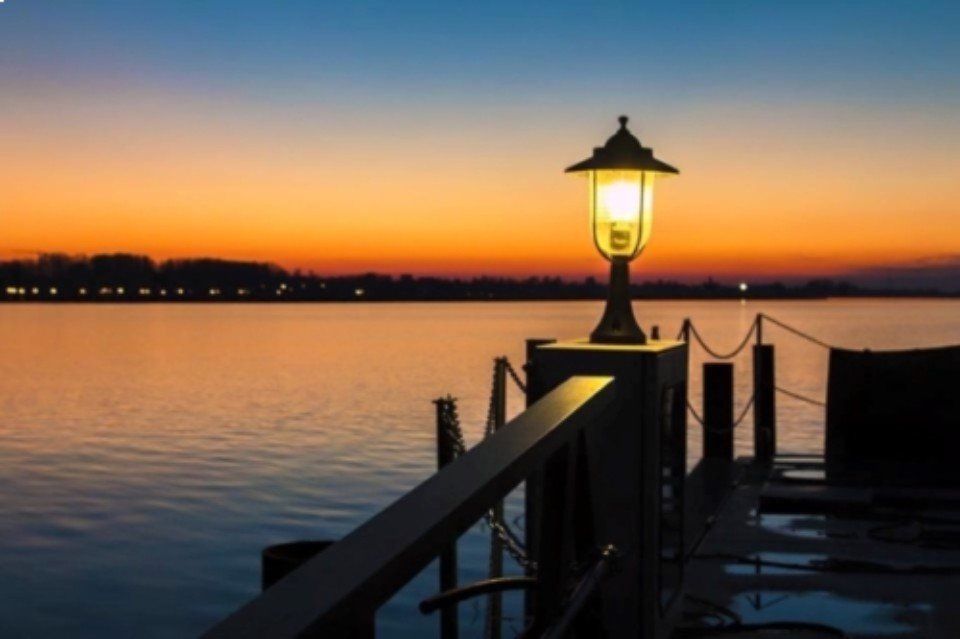 Una lanterna accesa illumina la superficie calma di un lago al tramonto, con un cielo arancione e blu.