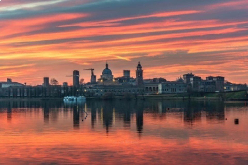 Il profilo della città al tramonto si riflette sull'acqua calma, sotto nuvole striate di arancione e rosa.