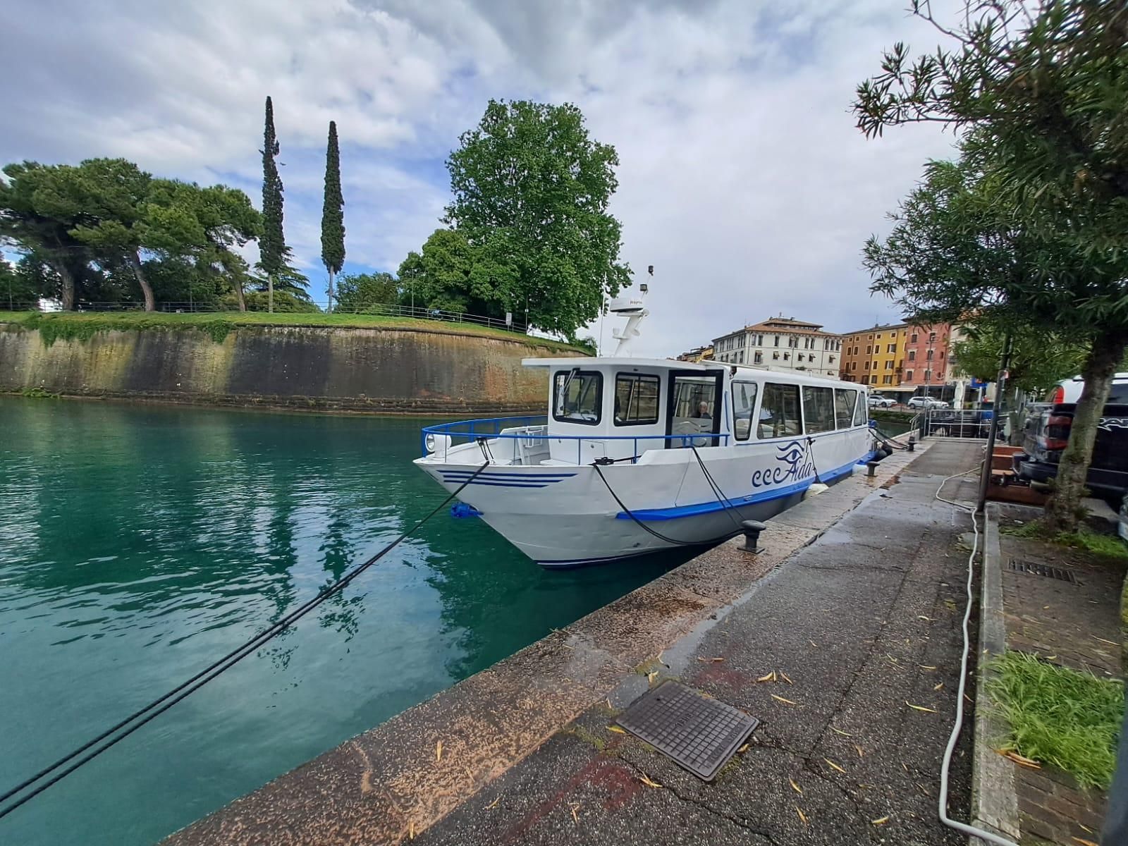 Barca bianca ormeggiata su un fiume turchese accanto a una passerella di pietra, con alberi ed edifici lungo la riva.