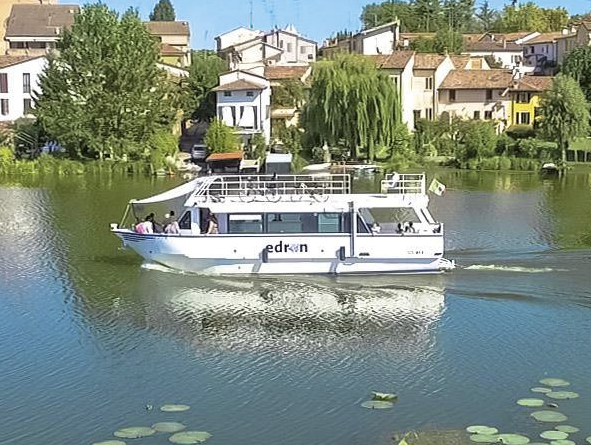 Barca turistica bianca su un lago tranquillo con case e alberi lungo la riva.