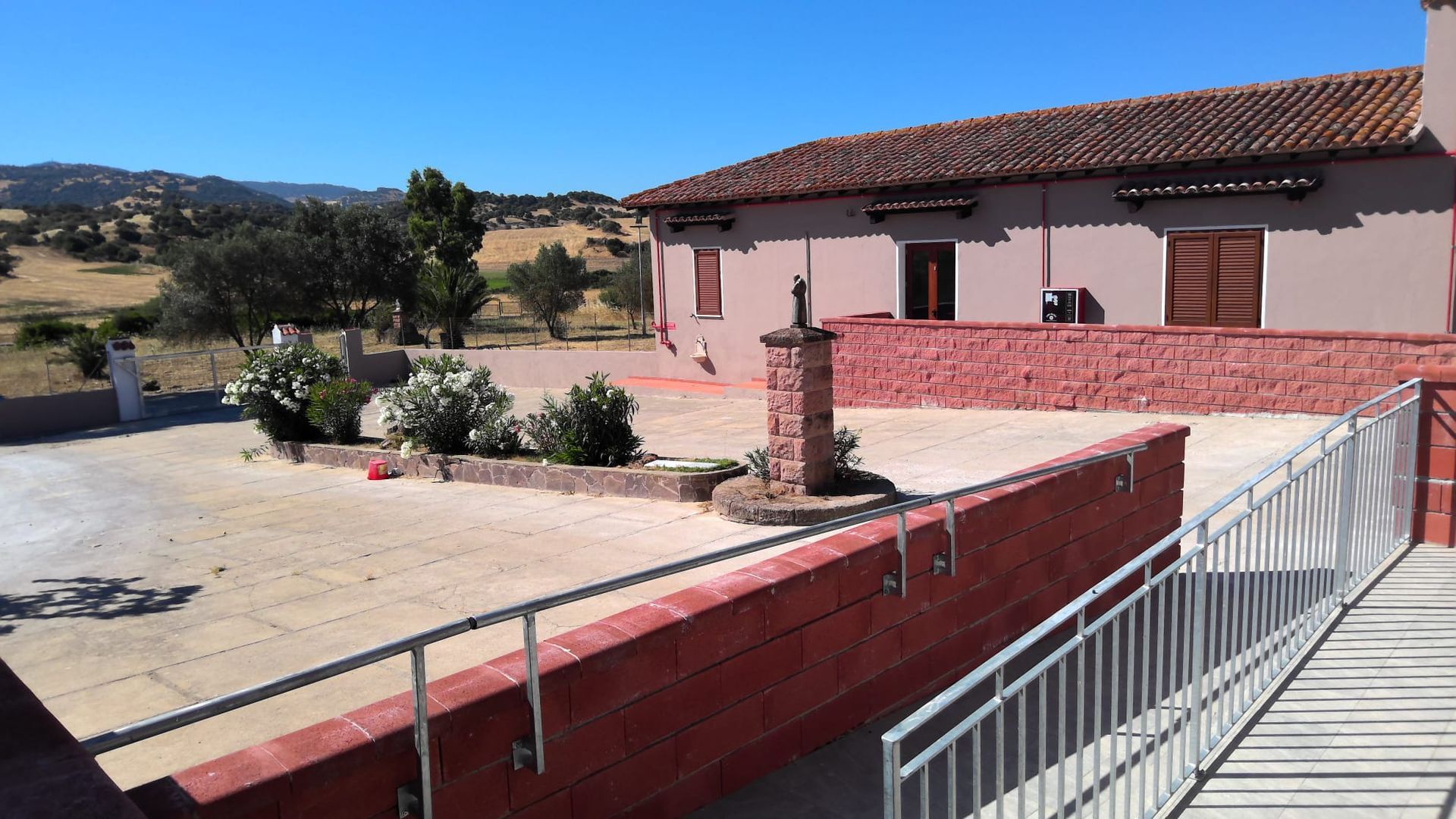 Casa rurale con pareti rosa, tetto di tegole rosse e recinzione bianca accanto a un cortile lastricato e soleggiato.