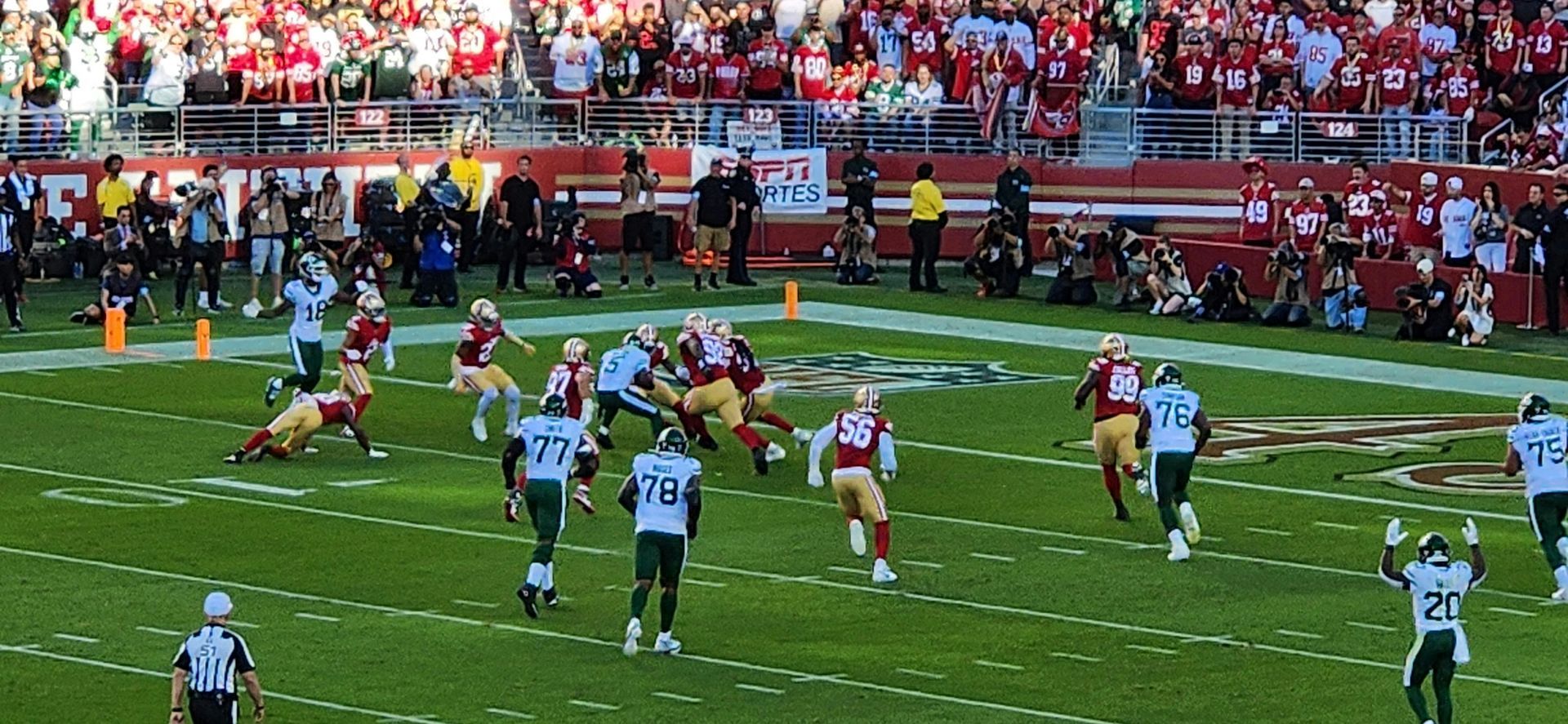 Football game, players in action on a green field, surrounded by stadium seating filled with red-clad fans.