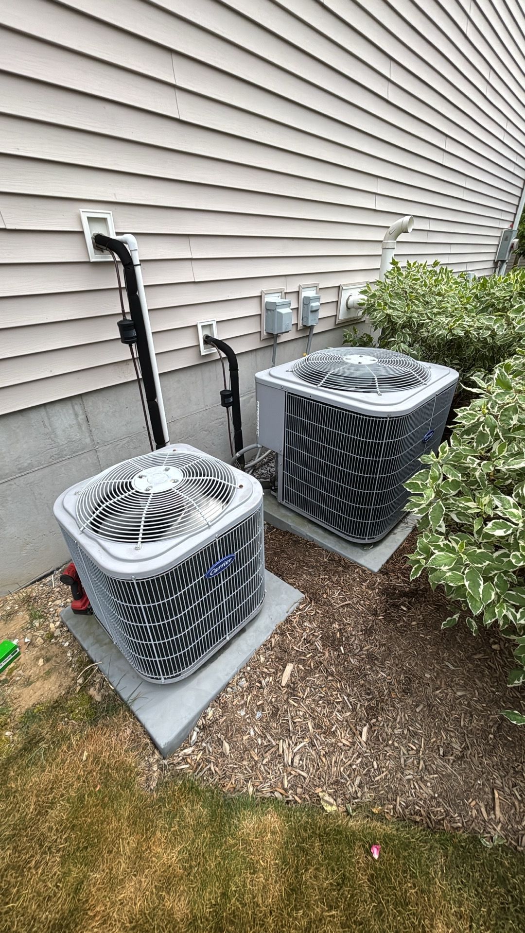 Two outdoor air conditioning units beside a house wall, next to bushes and grass.