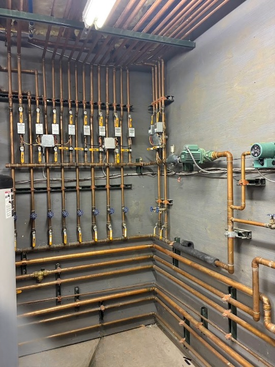 Industrial utility room with copper pipes, valves, and wiring on a gray wall and ceiling.