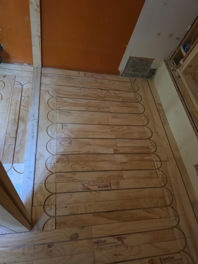 Floor heating mat laid on unfinished subfloor beside a brown door in a hallway.