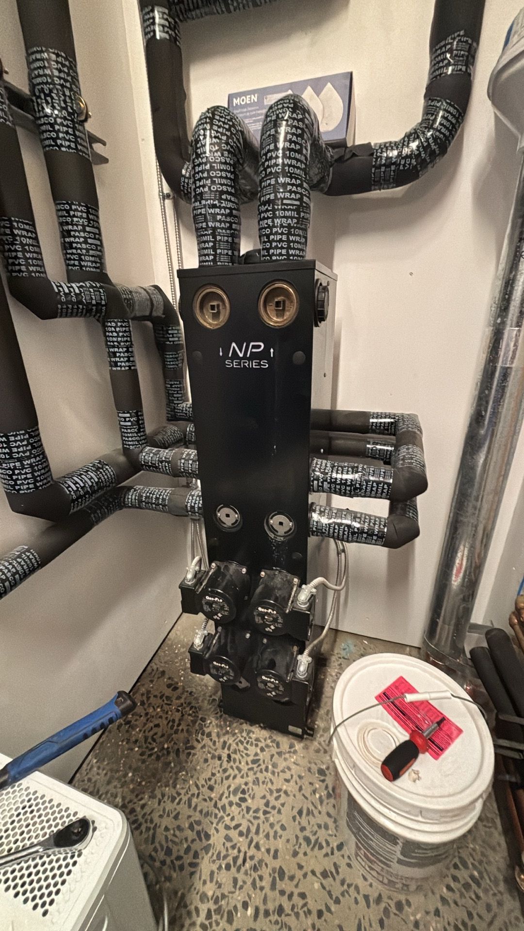 Black plumbing manifold with wrapped pipes in a utility closet, beside a white bucket on the floor