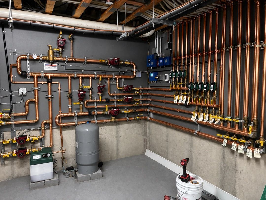 Utility room with copper pipes, valves, tanks, and wall-mounted plumbing equipment along concrete walls.