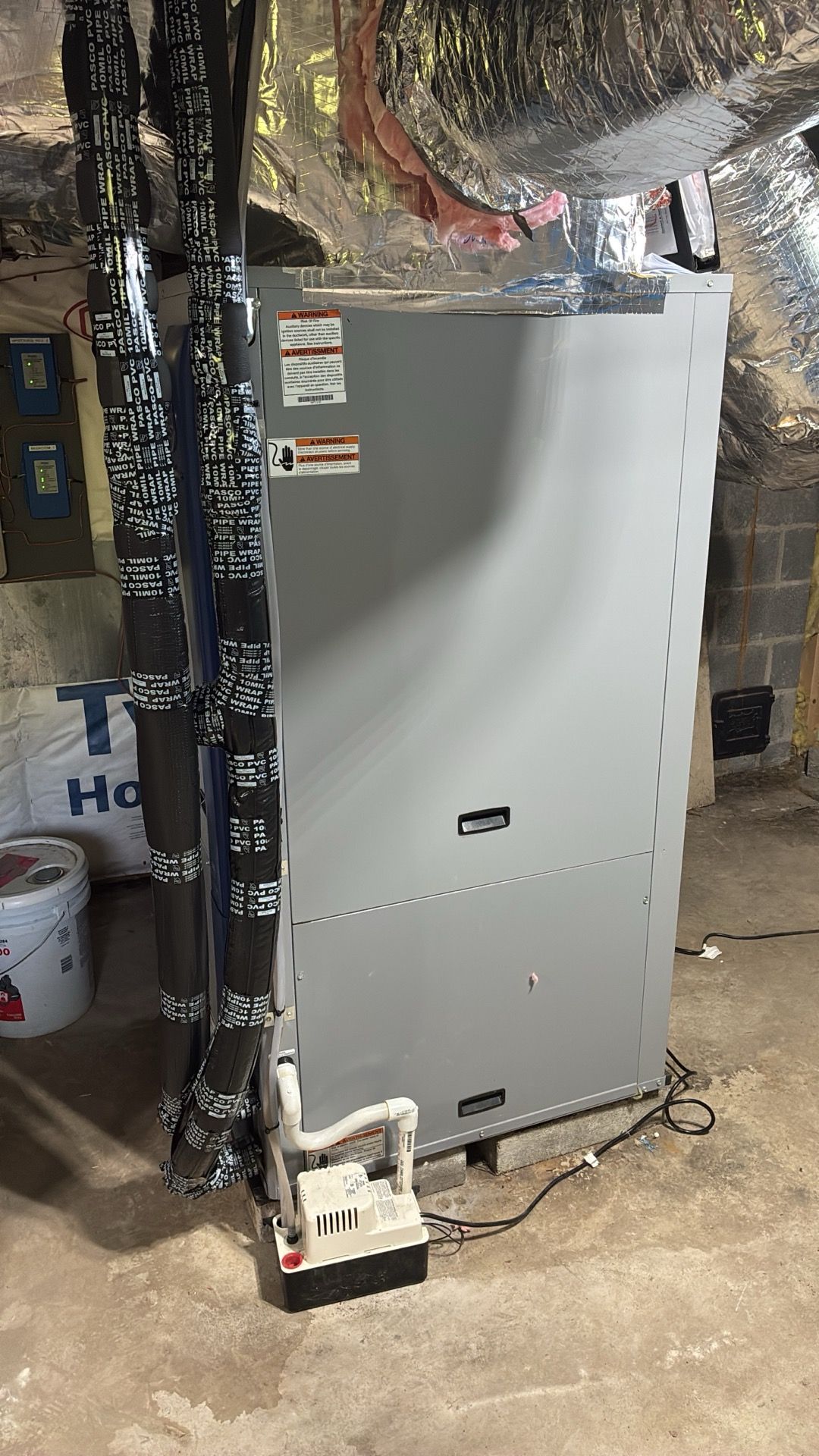 Gray HVAC unit in a basement or utility area with exposed pipes and duct insulation nearby