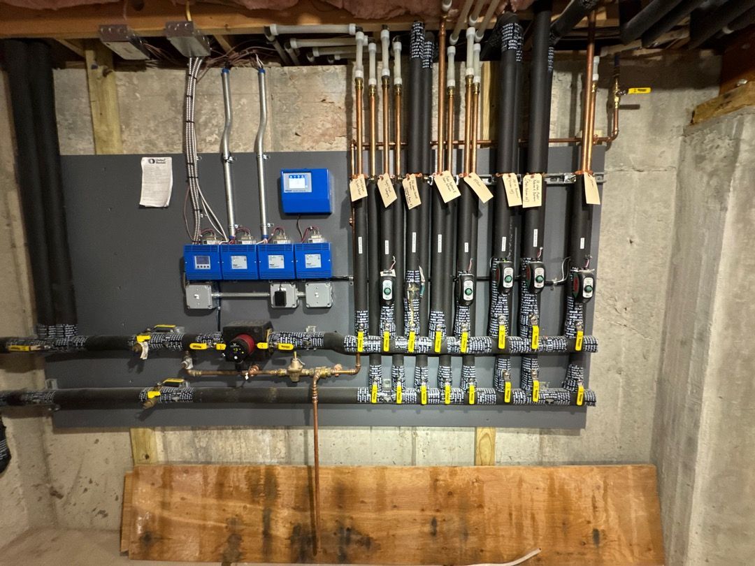 Basement utility wall with plumbing manifold, pipes, gauges, and blue control boxes above a wooden board.