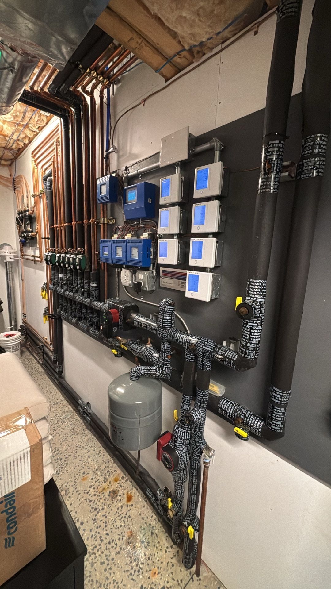 Wall-mounted industrial plumbing and electrical control panel with pipes, valves, and blue-labeled switches in a utility room