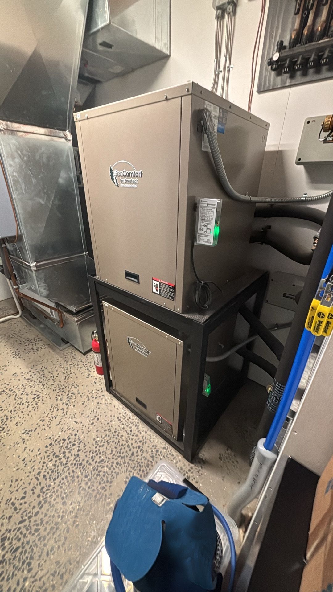 Gray HVAC equipment stacked in a mechanical room beside pipes and a blue chair
