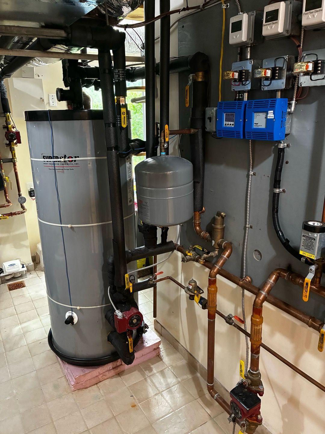 Boiler room with water heater, pipes, valves, and blue control boxes on a tiled floor.