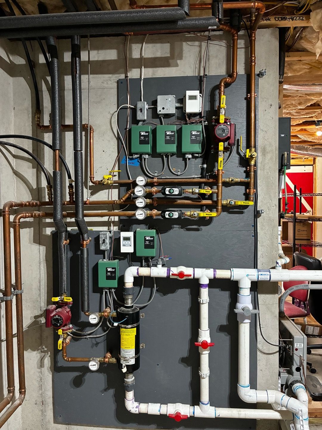 Industrial piping and control valves on a wall, with green pumps and white PVC pipes in a utility room