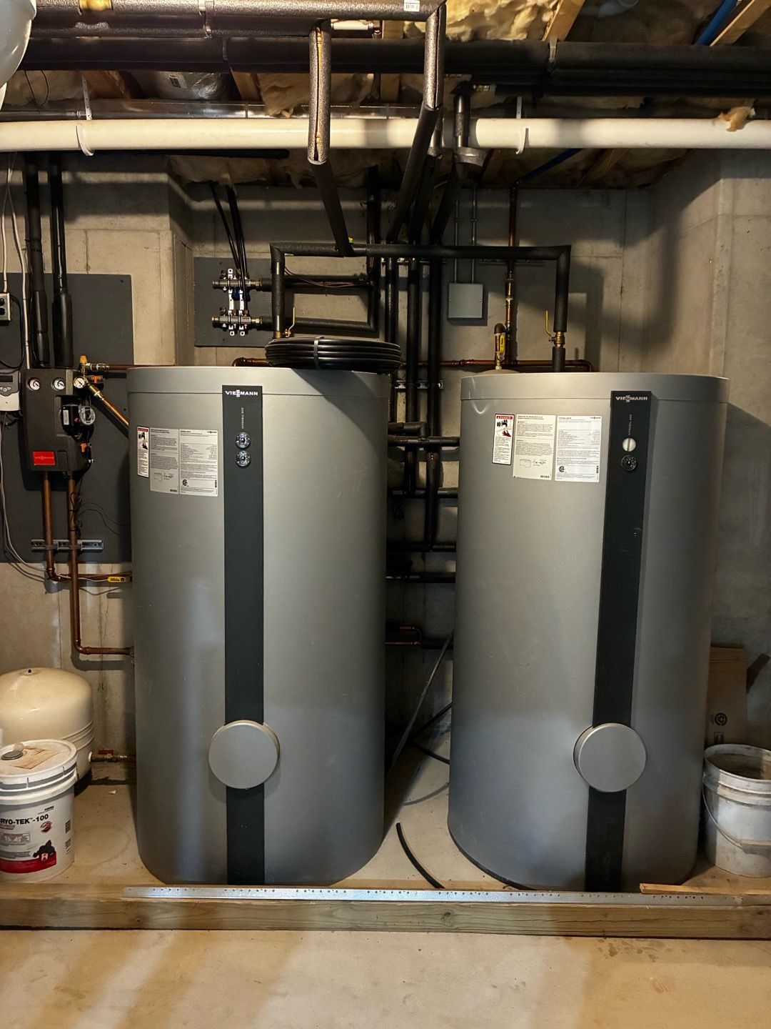 Two gray water heaters in a basement utility room with pipes and valves above them