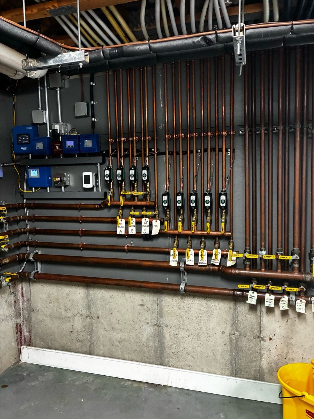 Basement utility wall with copper pipes, valves, gauges, control boxes, and a yellow bucket on the floor