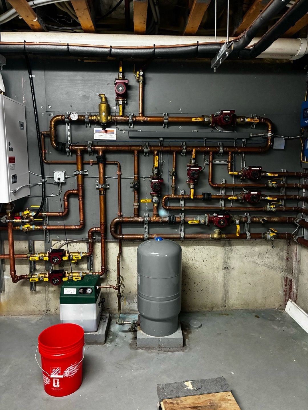 Basement utility room with exposed pipes, valves, a gray tank, and a red bucket on the floor