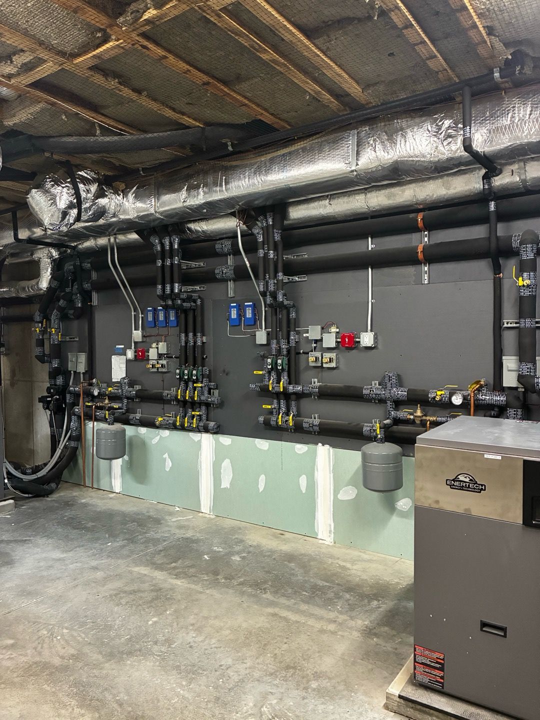 Industrial mechanical room with black pipes, valves, and insulated ducting along a gray wall